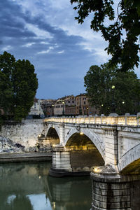 Illuminated bridge over river in town against cloudy sky