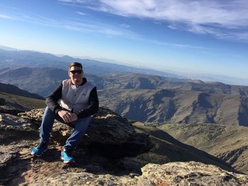 Portrait of young man sitting on rock against sky