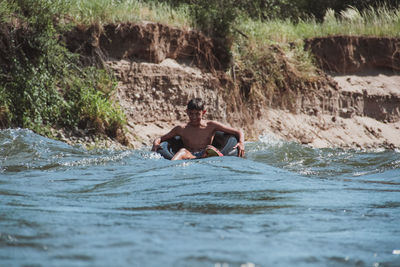 Full length of shirtless man sitting on rock in sea