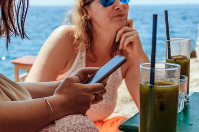 Close-up of woman sitting in beach bar, using mobile phone, drinking matcha latte