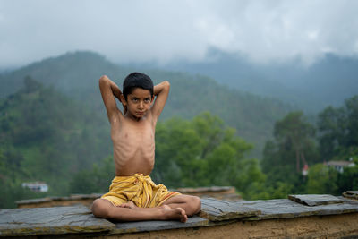 Midsection of shirtless man sitting on mountain against trees