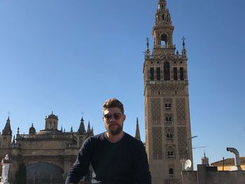 Portrait of young man wearing sunglasses standing against church in city during sunny day