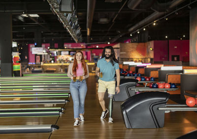 Happy man and woman with glasses of beer walking in bowling alley