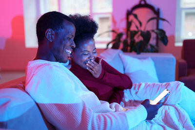 Happy african family couple laughing while relaxing on sofa and looking at smartphone screen