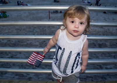 High angle portrait of baby girl holding american flag while standing by railing at beach