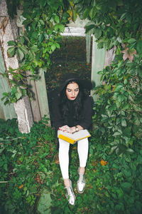 High angle view of young woman reading book while sitting amidst plants