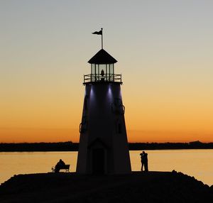 Silhouette people and lighthouse by sea against sky during sunset