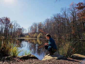 Side view of woman crouching by lake against sky in forest