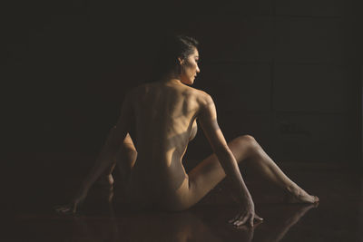 Rear view of naked woman looking away while sitting against black background