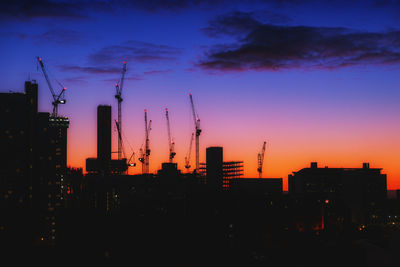 Silhouette cranes in city against sky during sunset