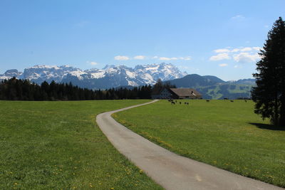 Empty road on grassy field by snow covered mountains against sky