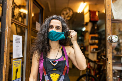 Positive adult ethnic female mechanic in colorful apron with protective mask smiling and looking at camera while standing on entrance of bike service workshop