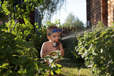 Happy little boy in watersport goggles with a pistol in hands watering the garden in summer cottage