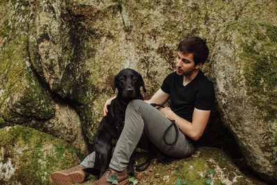 Young man with a black labrador retriever sitting on a mossy rock