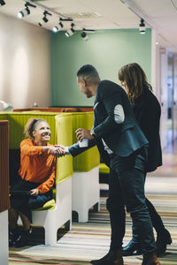 Smiling businesswoman sitting while shaking hands with businessman by female colleague standing in corridor at office