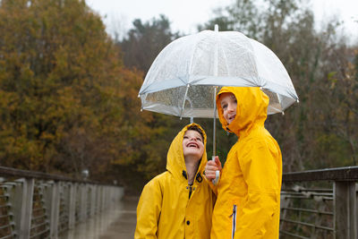 Smiling adorable siblings wearing yellow raincoats standing under umbrella on bridge in rainy day in autumn and enjoying weather