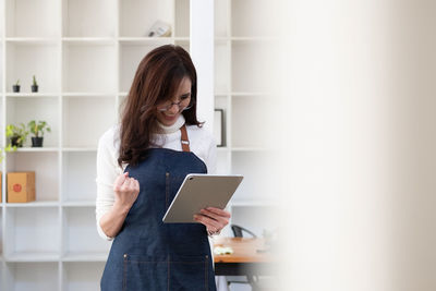 Young woman using digital tablet while standing against wall