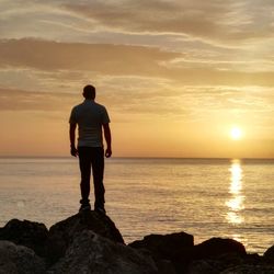 Full length rear view of man standing on rock against sea and sky during sunset