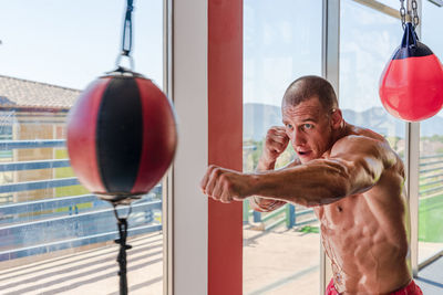 Side view of determined male boxer in boxing gloves punching heavy bag during intense workout in gym