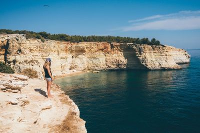 Rear view of man standing on cliff by sea against sky