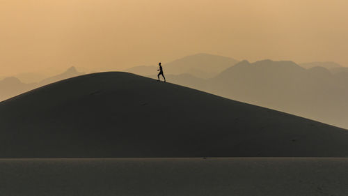 Silhouette person walking on mountain by sea against sky during sunset