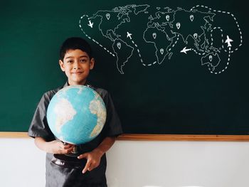 Portrait of boy holding globe while standing against blackboard in classroom