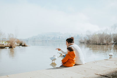 Mother with daughter sitting at lakeshore against sky