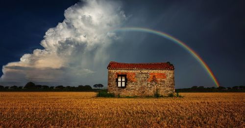 House on field against rainbow in cloudy sky