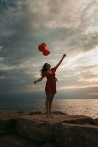 Woman holding balloons while standing on rock by sea against sky during sunset