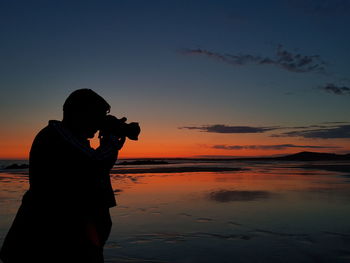 Silhouette man photographing sea against sky during sunset