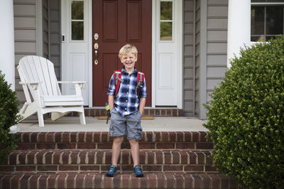Grinning blonde boy with hands in pockets stands on brick front steps