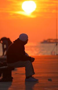 Silhouette man sitting by sea against orange sky
