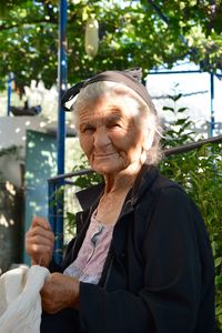 Portrait of senior woman sewing plastic while standing against fence