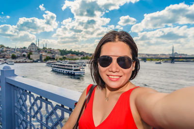 Portrait of smiling woman wearing sunglasses taking selfie while standing by railing against sky