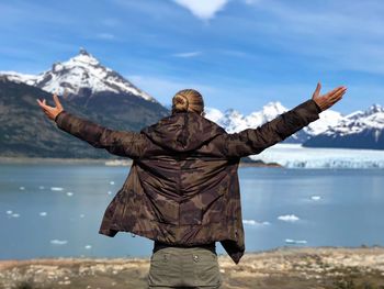 Rear view of man with arms outstretched standing against lake during winter