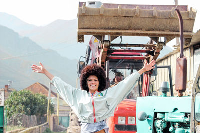 Portrait of happy young woman with arms outstretched standing against bulldozer