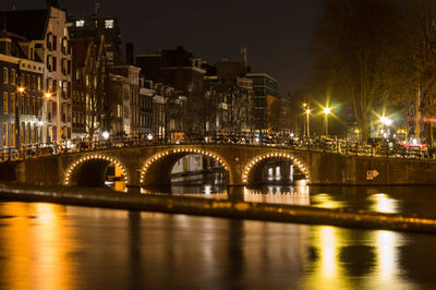 Illuminated bridge over river by buildings against sky at night