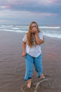 Delighted young curvy female standing barefoot on wet beach near sea and having fun while looking at camera and touching face