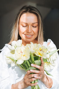 Young smiling woman with bouquet of yellow flowers in hands near window with hard light at home