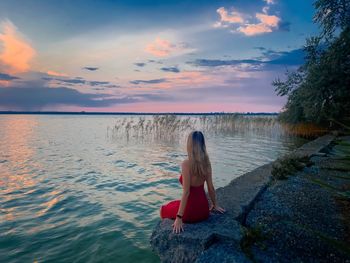 Rear view of woman in red dress sitting down near the lake at sunset and looking at the sky