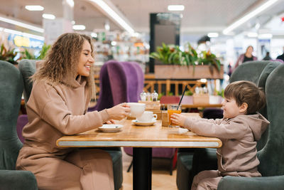 Young curly haired mother and her little son have breakfast or lunch in a cafe, sitting at a table