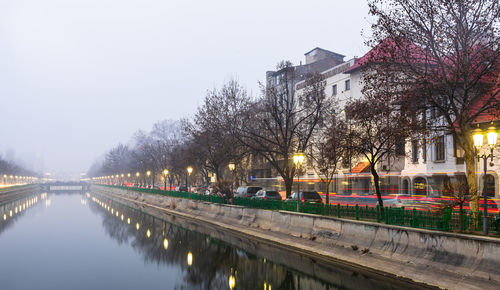 Illuminated street by canal against sky at dusk