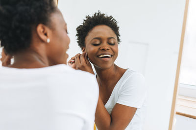 Portrait of happy young african american woman in white t-shirt puts on an earring near mirror