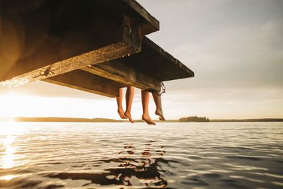 Low section of female friends sitting on jetty by lake during sunset