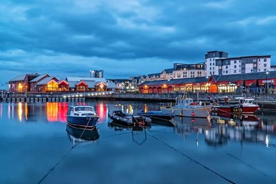 Boats moored on river against illuminated buildings in city at dusk