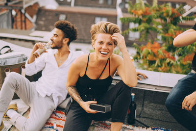 Cheerful woman enjoying music on speaker while sitting with friends on terrace at party