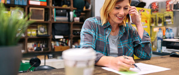 Portrait of smiling blonde young woman receiving order by phone for ecommerce shop