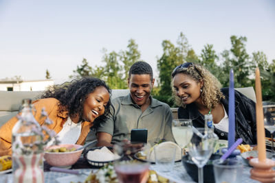 Smiling man sharing smart phone with female friends during dinner party in back yard