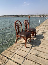 Empty chairs and table on pier by sea against sky