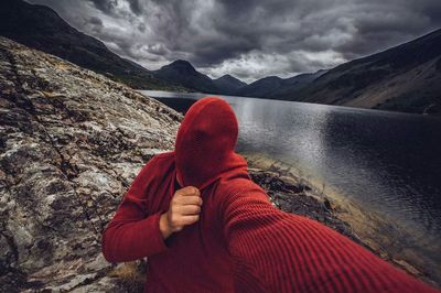 Man with face covered with sweater by lake against mountains and cloudy sky